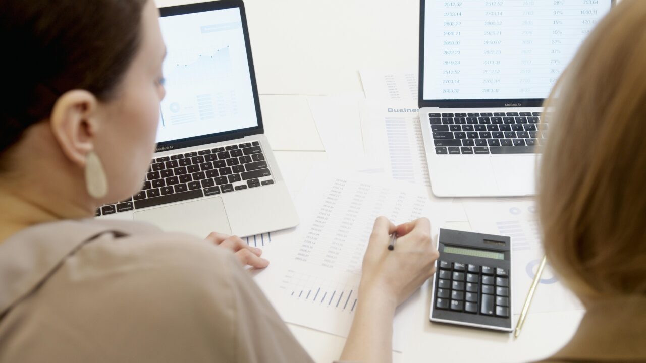 Two professionals at a desk filled with spreadsheets and data charts, looking at two laptops and using a calculator to analyze financial information.