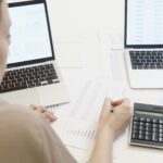 
                            Two professionals at a desk filled with spreadsheets and data charts, looking at two laptops and using a calculator to analyze financial information.                            