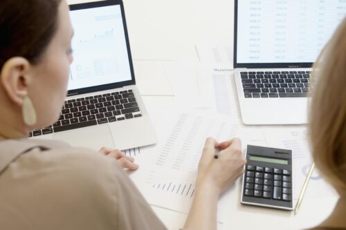 
                Two professionals at a desk filled with spreadsheets and data charts, looking at two laptops and using a calculator to analyze financial information.